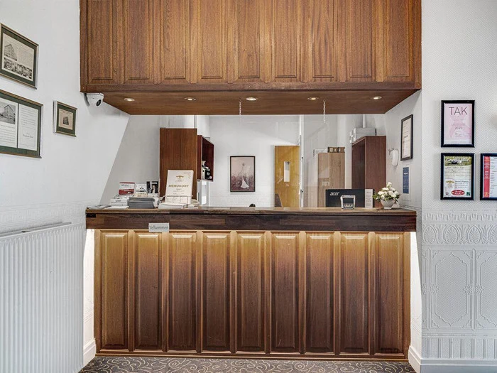 En hotelreception med træpaneler, priser og certifikater på væggene, en indskrivningsbog og et lille blomsterarrangement på skranken.