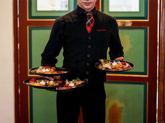 En tjener i sort vest og rødternet slips bærer fire tallerkener med forskelligt mad foran grønne og brune døre.