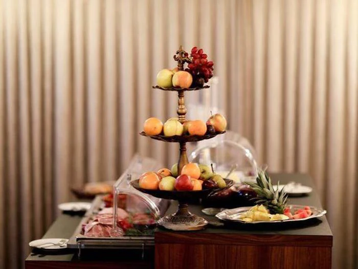 Et frugtstativ med æbler og vindruer og en bakke med forskellige frugter på et buffetbord med overdækkede fade foran beige gardiner.