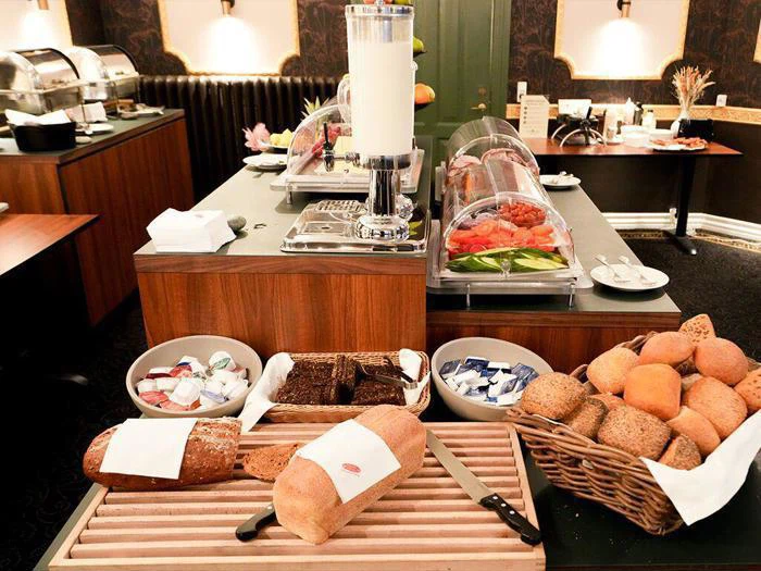 En morgenmadsbuffet med forskellige brød, rundstykker, smørportioner og pålæg på træborde i et godt oplyst spiseområde.