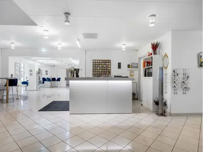 Moderne, lyst receptionsområde med hvid skranke, klinkegulv og minimal indretning med planter, vægkunst og reoler. Blå stole ses i baggrunden.