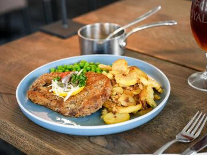 En blå tallerken med en paneret kødkotelet toppet med citron og krydderurter, sprøde kartofler og grønne ærter, serveret på et træbord.