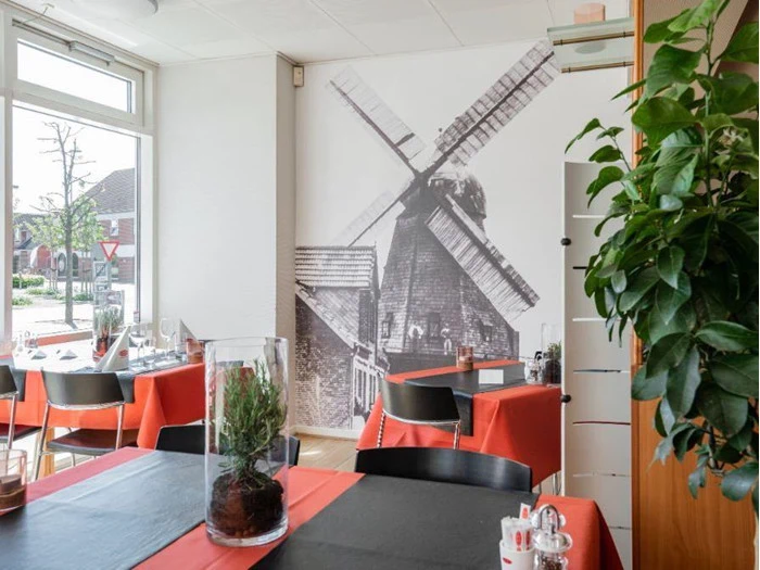 En hyggelig restaurant med røde duge, sorte stole og et stort vægmaleri af en vindmølle. På bordene står en plante og en glasdekoration, og sollyset strømmer ind gennem vinduet.