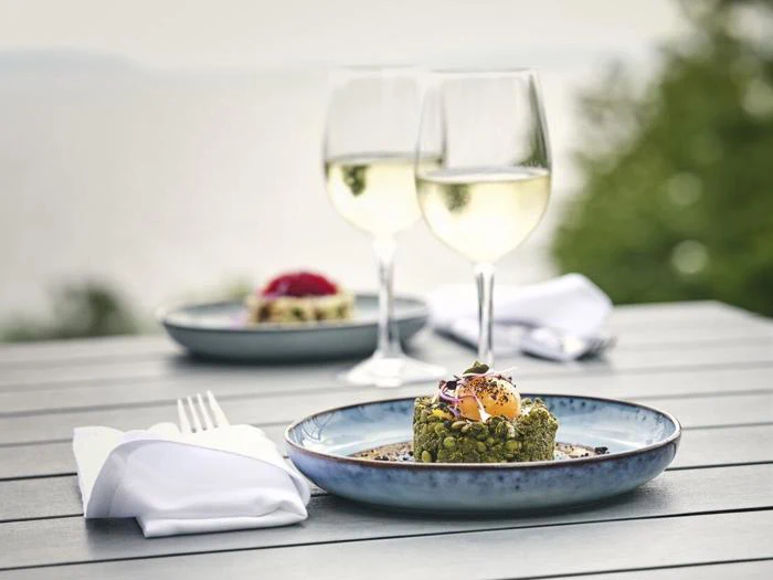 En gourmetret med gul topping på en blå tallerken, to glas hvidvin og en anden desserttallerken på et udendørs bord med en sløret naturlig baggrund.