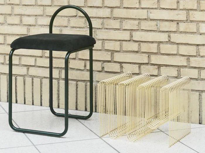 En moderne sort stol med afrundet ryglæn står ved siden af tre skulpturelle former i guldmetal på et flisegulv op ad en murstensvæg.