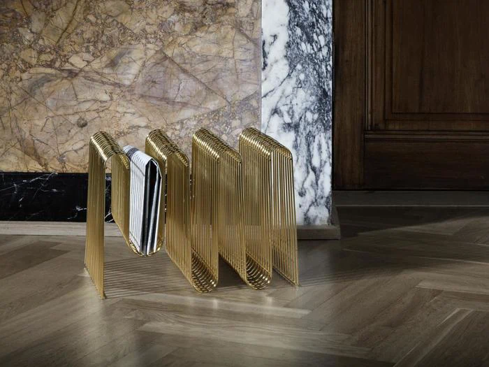 En magasinholder i guldmetal med magasiner, placeret på et trægulv ved siden af en væg med brune og hvide marmormønstre.