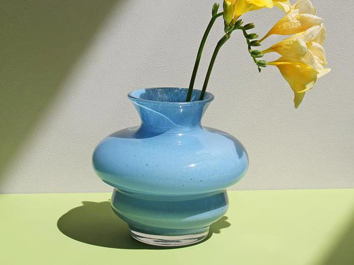 En skinnende lyseblå vase med to gule blomster står på en lysegrøn flade og kaster en blød skygge mod en lys væg.