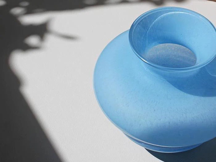 En gennemskinnelig blå vase står på en hvid flade, delvist i sollys og skygge, med slørede bladskygger i baggrunden.