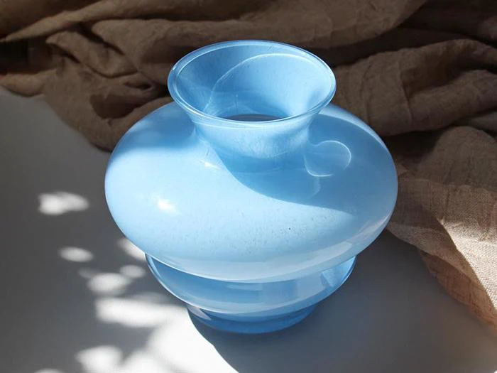 En blank, lyseblå glasvase med en bred, buet krop og en smal åbning står på en hvid flade med et beige stof i baggrunden.