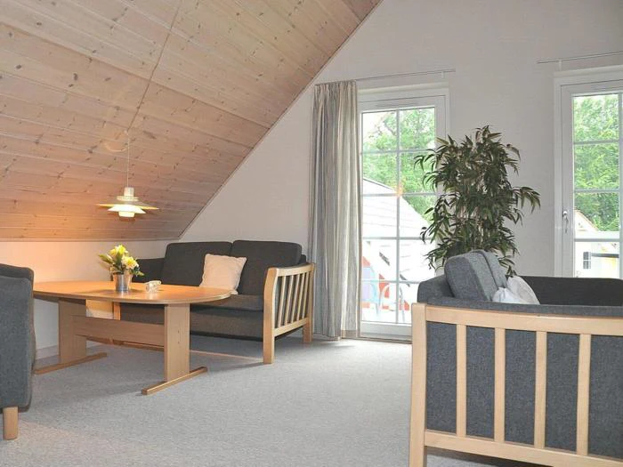 En hyggelig stue med skråt træloft, to sofaer, et rundt sofabord i træ med blomster, en høj plante og store vinduer, der lukker naturligt lys ind.