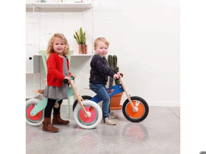 To små børn, en pige i en rød cardigan og en dreng i en marineblå jakke, kører på farverige løbecykler indendørs med planter og hvide hylder i baggrunden.