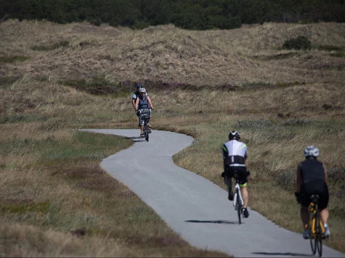 Tre cyklister kører ad en snoet asfalteret sti gennem græsklædte, bølgende bakker under en klar himmel.