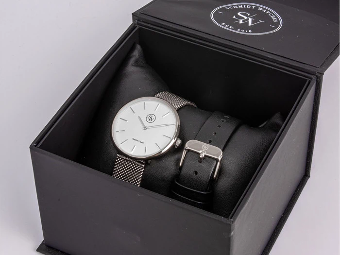 Et sølvfarvet Schmidt-armbåndsur med en metalnetrem og en ekstra sort læderrem i en åben sort gaveæske.
