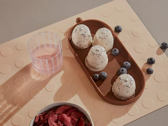 Et glas vand, en bakke med tre hvide, birkesdækkede is og flere blåbær på en beige måtte med hævede cirkler. En skål med tørret frugt er delvist synlig.