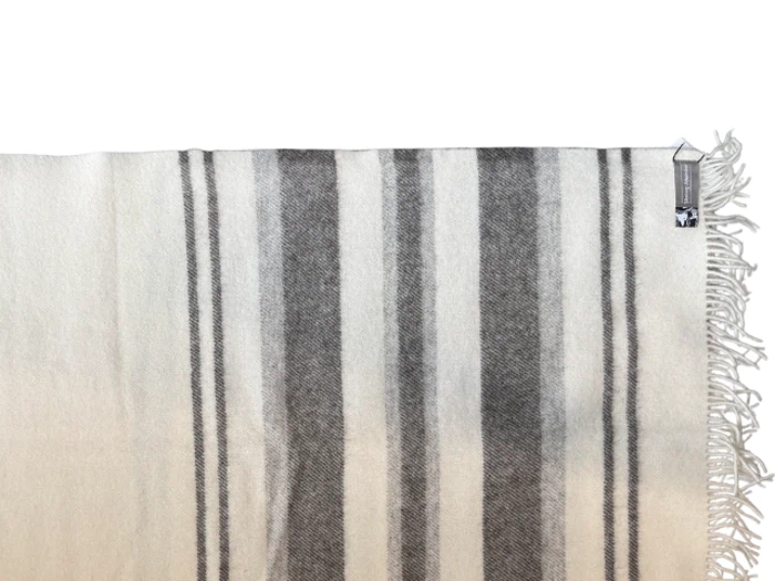 Et uldtæppe med sorte og grå lodrette striber og frynser på den ene kant hænger på en hvid vægknage mod en ensfarvet baggrund.