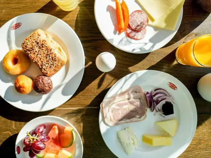 Morgenmad med brød, kager, ost, pålæg, skiveskårne grøntsager, frugt, vindruer og glas med appelsinjuice arrangeret på et træbord.