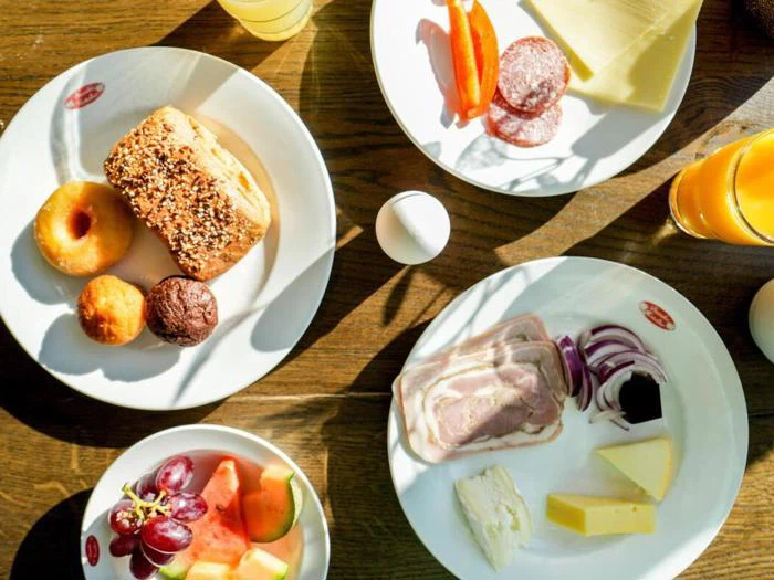 Fire tallerkener med forskellige morgenmadsprodukter: kager, donuts, muffins, oste, pålæg, frisk frugt, vindruer, appelsinjuice og et hårdkogt æg på et træbord.