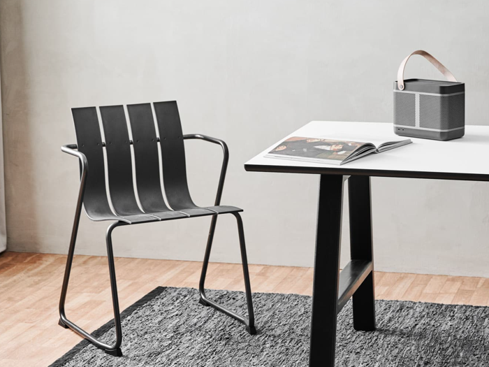 Et minimalistisk rum med en moderne sort stol, et hvidt bord med sorte ben, et åbent magasin og en bærbar højttaler med læderrem på et gråt tæppe og trægulv.