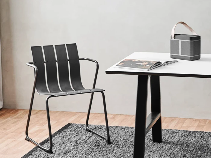 Et minimalistisk rum med en moderne sort stol, et hvidt bord med sorte ben, et åbent magasin og en bærbar højttaler med læderrem på et gråt tæppe og trægulv.