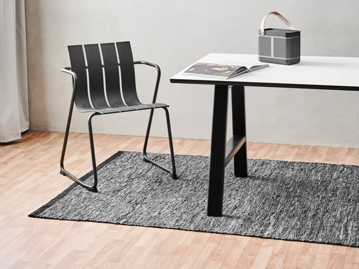 En moderne sort stol og et hvidt bord står på et gråt tæppe i et minimalistisk rum. På bordet ligger en åben bog og en lille højttaler med læderrem.