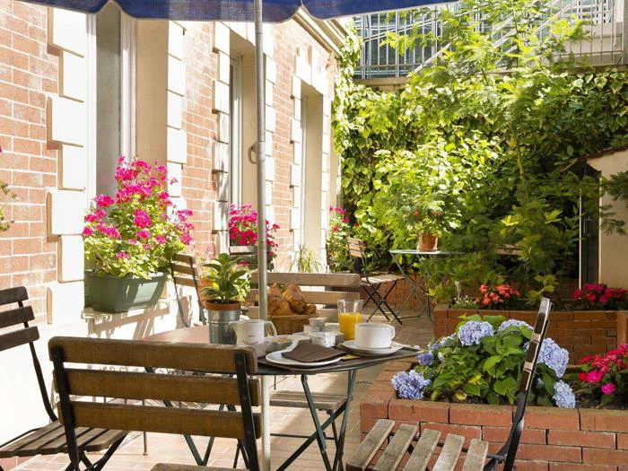 En hyggelig udendørs terrasse med træstole og -borde, morgenmadsprodukter, farverige blomster, frodigt grønt og murstensvægge i baggrunden.