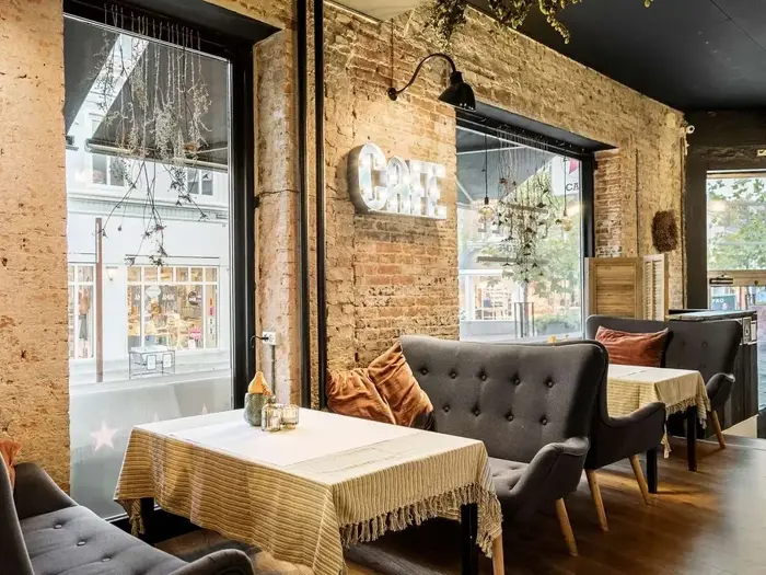Et hyggeligt caféinteriør med murstensvægge, store vinduer, grå, tuftede sofaer, borde med duge og varm, dekorativ belysning.