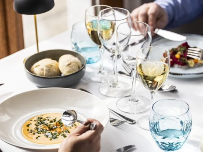 En person sidder ved et bord og spiser suppe af en hvid skål, med vinglas, en skål med brød og en tallerken med mad ved siden af.