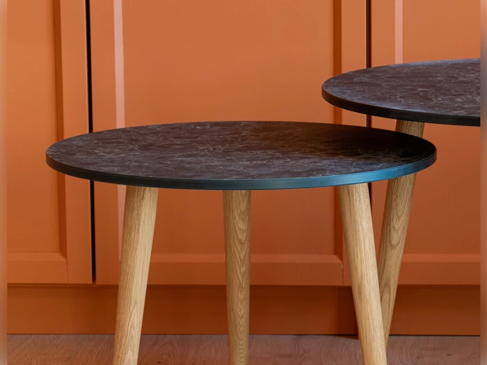 To runde borde med mørke plader og lyse træben, opstillet mod en væg med orange paneler og et trægulv.