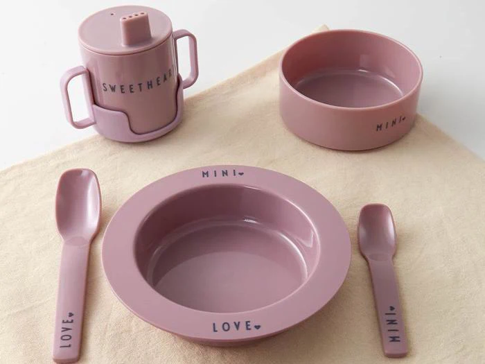 Et sæt lyserødt børneservice med skål, tallerken, kop med låg, gaffel og ske, hver især mærket med ord som MINI, LOVE og SWEETHEART, arrangeret på et beige klæde.