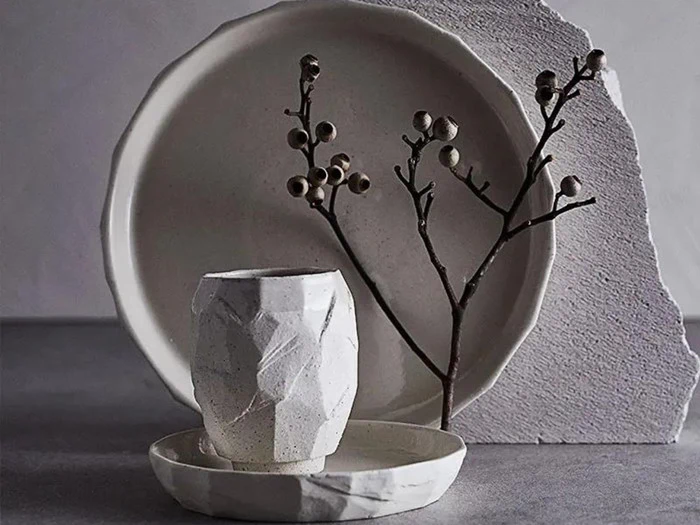 Minimalistiske keramiktallerkener og en facetteret kop i hvid, arrangeret med en tynd tørret gren med små runde bær på en struktureret baggrund.