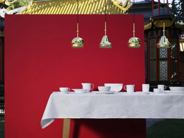 En hvid dug med hvide kopper og skåle er dækket foran en rød væg med fire guldpendler over og en traditionel asiatisk bygning i baggrunden.
