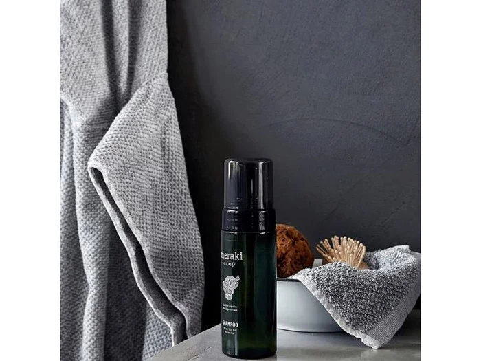 En mørkegrøn Meraki sæbedispenser står på en grå overflade ved siden af en hvid skål med et håndklæde, en svamp og en pind som dekoration, og i baggrunden hænger et gråt håndklæde.