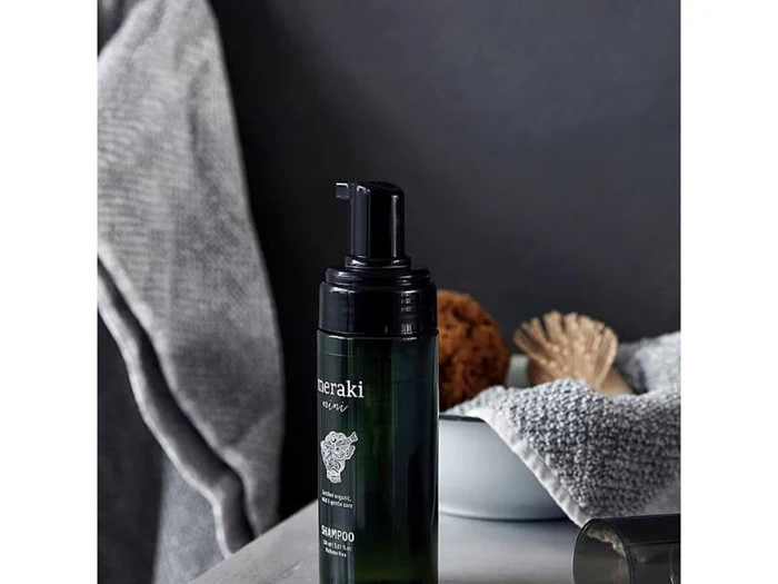 En sort pumpeflaske med shampoo står på et badeværelsesbord ved siden af en skål med en svamp og en muslingeskal, og et gråt håndklæde er hængt op i nærheden.