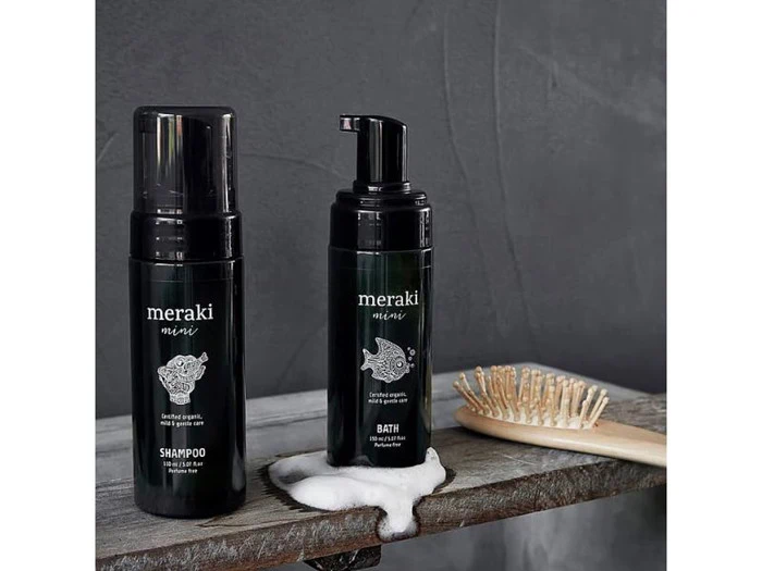 To sorte flasker med Meraki Mini badeprodukter og en hårbørste af træ står på en rustik hylde; skummet er synligt nær flaskerne mod en grå baggrund.