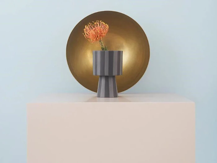En enkelt orange blomst i en moderne, stribet grå vase står på en beige piedestal med en stor, rund guldskive i baggrunden mod en lyseblå væg.