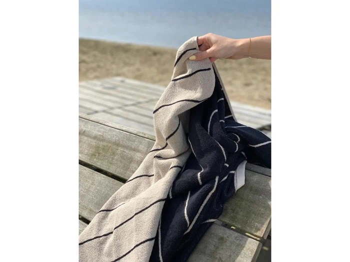 En hånd holder et sort- og hvidstribet håndklæde, der er draperet over en træbænk nær en sandstrand med vand i baggrunden.