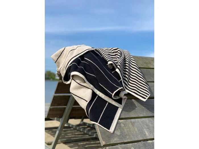 Et sort- og hvidstribet håndklæde er draperet over en træbænk ved en søstrand under en klar blå himmel.