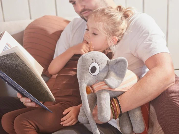 En mand læser en bog for en ung pige, der sidder på hans skød med en grå tøjelefant i hånden, mens de begge slapper af i en sofa.