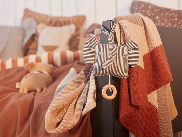 Et hyggeligt børneværelse med en elefant i plys, der hænger på en tremmeseng, et stribet tæppe og en blød, brun seng med dyrepuder i baggrunden.