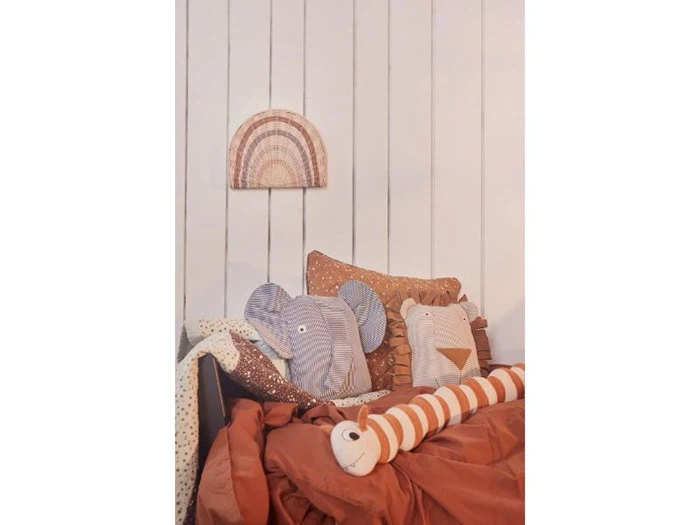 En hyggelig seng med rustfarvet sengetøj, dyreformede puder, et polkaprikket tæppe og en vævet regnbuevægdekoration over hovedgærdet.