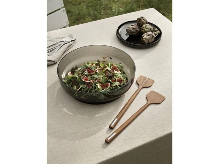 En skål med frisk salat med figner og grønt står på en beige dug, ledsaget af salatbestik i træ og en tallerken med to artiskokker i baggrunden.