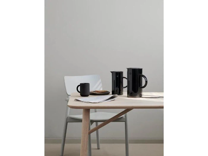 Et lyst træbord med sorte krus, sorte kander og en tallerken ved siden af en lysegrå stol mod en almindelig grå væg.