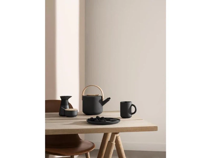 Et minimalt træbord med en sort tekande, et krus, en lille kande og en tallerken med bær ved siden af en brun stol i et svagt oplyst rum.