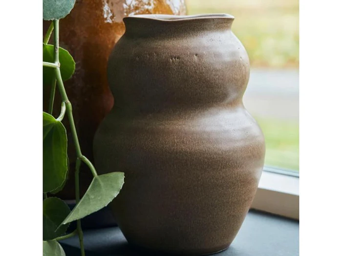 En brun, ujævnt formet keramikvase står i vindueskarmen ved siden af en grøn slyngplante med en sløret baggrund udenfor.