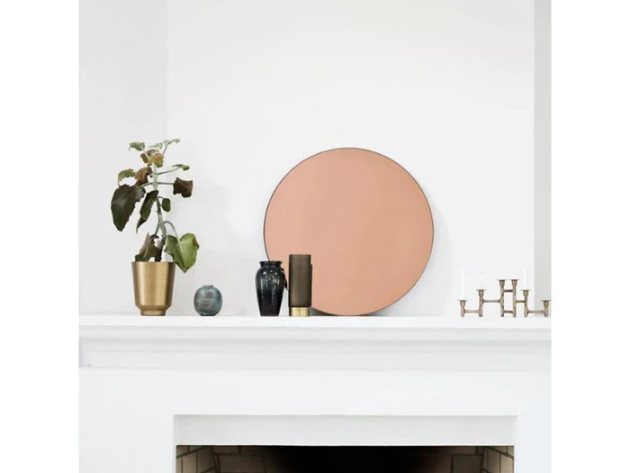 En minimalistisk kamin med en potteplante, et rundt ferskenfarvet spejl, to små vaser og en guldlysestage arrangeret på en hvid flade.