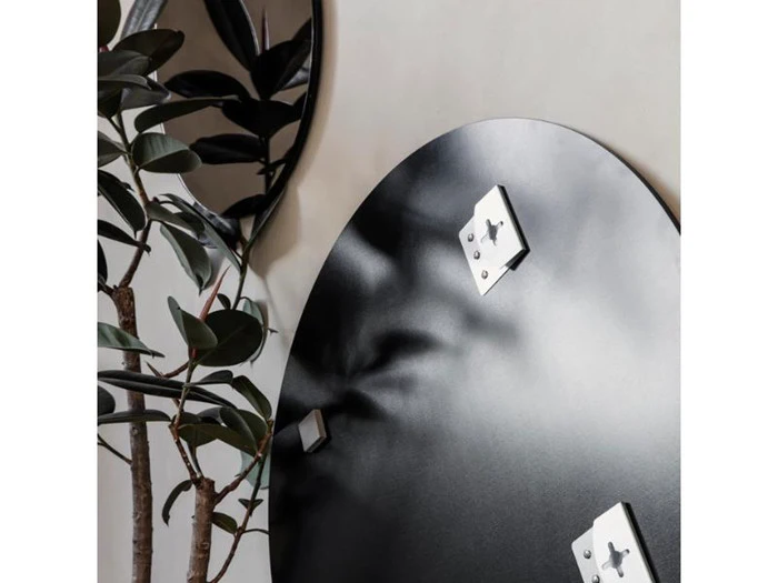 Et nærbillede af et rundt sort vægspejl med metalbeslag ved siden af grønne grene, der reflekterer lyset og en del af bladene.