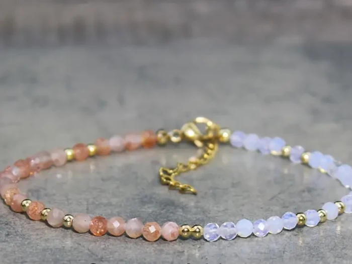 Et perlearmbånd med små facetterede perler i graduerede nuancer fra ferskenorange til lys lavendel, fremhævet med guldafstandsperler og en guldlås, vist på en grå overflade.