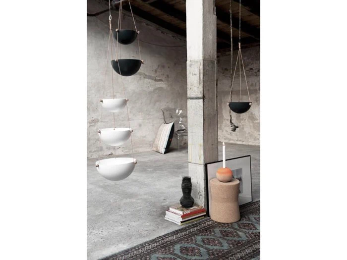 Hængende keramikskåle, hvide og sorte, i et minimalistisk betonrum med tæpper, bøger, en vase, kunstværker og en skammel med et stearinlys.