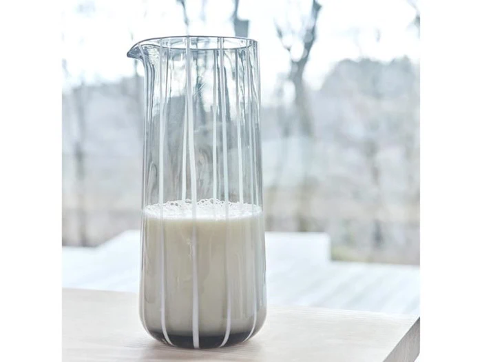 En klar glaskande med en lille mængde mælk står på en lys træoverflade med en sløret udendørs baggrund.