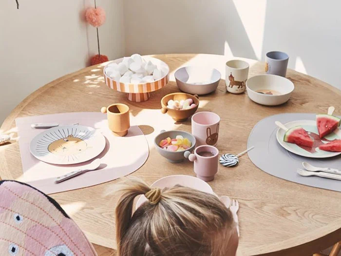 Et barn sidder ved et rundt træbord med tallerkener med vandmelon, skåle med snacks, kopper og farverigt service i stærkt sollys.